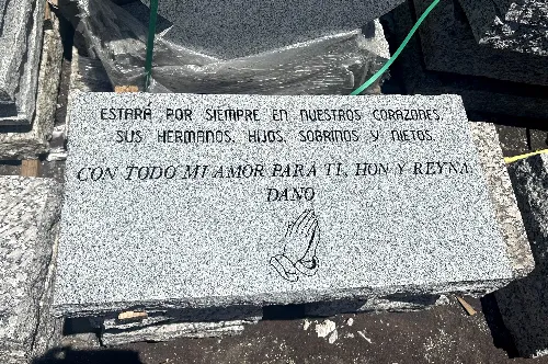 A gray granite memorial plaque with inscriptions in Spanish. It reads: "Estará por siempre en nuestros corazones, sus hermanos, hijos, sobrinos y nietos. Con todo mi amor para ti, hon y rey. Dano." There's an engraved image of praying hands at the bottom.