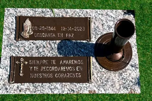 A gravestone with two plaques on a grassy area. The top plaque has dates and the phrase "Descansa en paz" with praying hands. The bottom plaque features a cross and reads "Siempre te amaremos y te recordaremos en nuestros corazones.