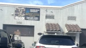 A small industrial building with a sign for "US Monument Memorials" featuring manufacturing services. Several cars are parked in front, including a white Honda. The building is light gray with two small awnings over the windows.