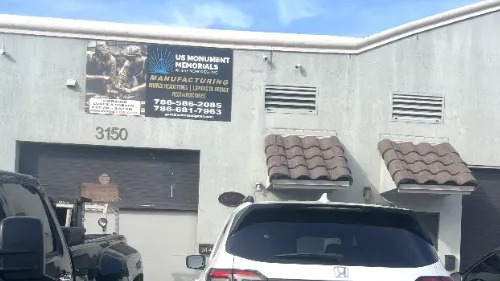 A small industrial building with a sign for "US Monument Memorials" featuring manufacturing services. Several cars are parked in front, including a white Honda. The building is light gray with two small awnings over the windows.