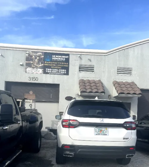 A white SUV and a black pickup truck are parked in front of a light gray industrial building with signage for "US Monument Memorials Manufacturing" above the entrance. The building has small windows and the license plate on the SUV is visible.